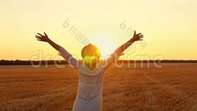 日落时，一个穿着白色连衣裙和草帽的女孩在麦田里举手。 一位年轻女子站着